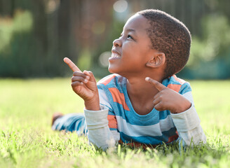 Boy, excited and pointing in park on grass for summer, development and weekend with sunshine. Black child, happy and fingers in nature for school vacation, relax and fun childhood with smile in Kenya