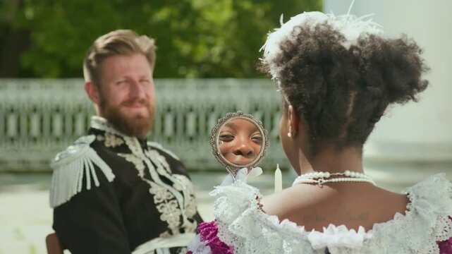 Young Black lady in feathered fascinator and victorian dress looking at her reflection in hand mirror and talking with Caucasian military officer in elaborate uniform while sitting outdoors