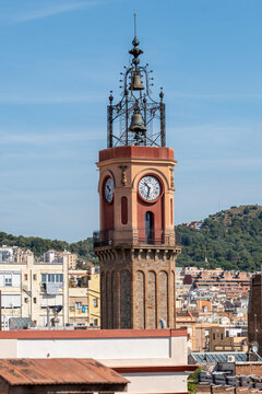 Featuring an iconic clock tower standing tall amidst an urban setting with buildings and greenery in the background, this image showcases a blend of historic and modern elements in Gracia Barcelona