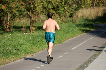 Man in shorts running on sidewalk along the city street, rear view. Runner in park, workout in autumn season