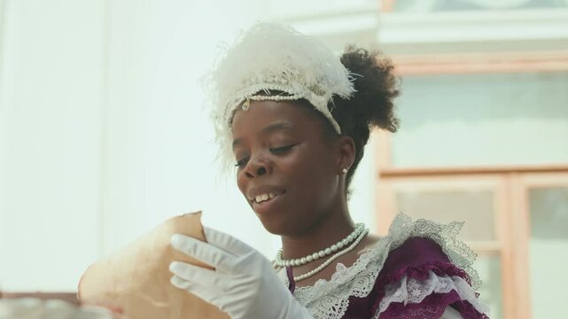 Young aristocratic African American lady wearing feathered fascinator, white gloves and period dress sitting outside on sunny day and reading romantic letter with pleasure