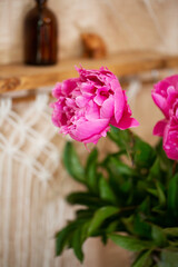 A bouquet of pink peonies with green leaves in a crystal vase on the background of a wooden shelf and macrame weaving.