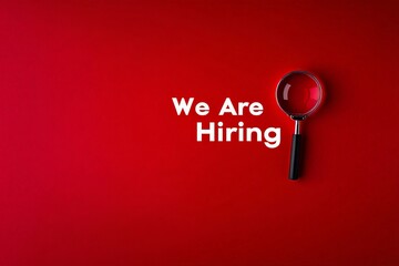 A clear 'We Are Hiring' sign on a red backdrop featuring a magnifying glass, emphasizing the active search for new employees and creating an inviting and urgent call to action.