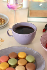 Plate of pastel macarons, cookies and chocolate, cup of tea of coffee, glass of bubble water, various berries, books and accessories on the table. Selective focus, pastel colors.