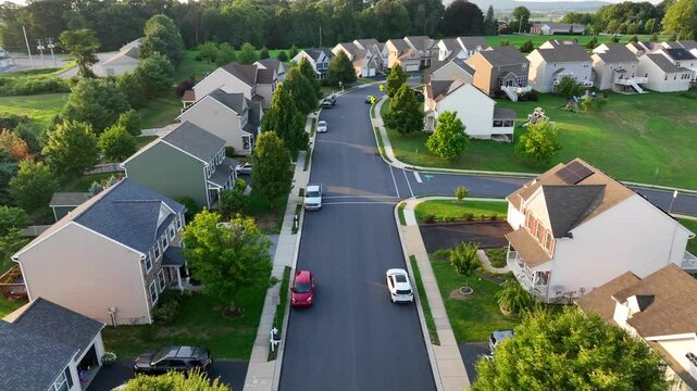 A quiet suburban neighborhood with modern homes and tree-lined streets in Pennsylvania. Curved roads, parked cars, and playgrounds visible in backyards. Aerial during late afternoon.