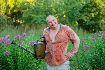 A man holds a watering can and smiles. He stands in a garden with flowers. Taking care of the garden.