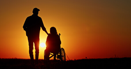 Obraz premium Silhouettes of a man and a woman in a wheelchair admiring a scenic sunset over a field.