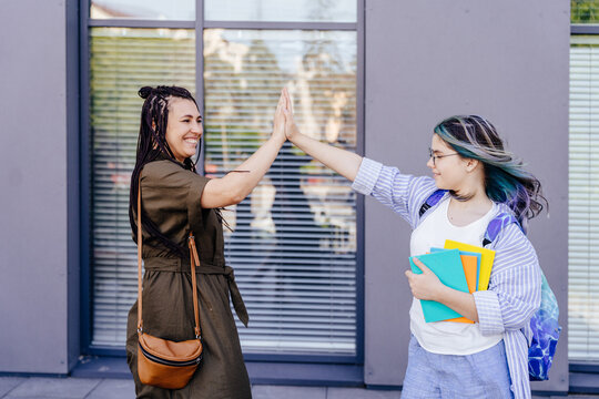 Mother and daughter giving high five in front of school. Mother take her daughter to school. Little girl tells mom goodbye. First day of school.
