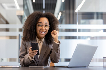 Portrait of young African American businesswoman who received good news and result, sitting in office at desk, holding phone, raised victory fist up and looking smiling at camera