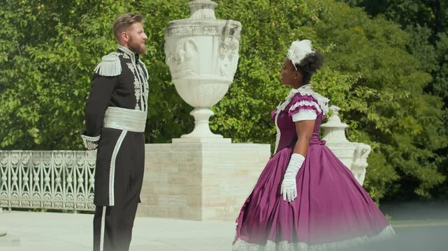 Black aristocratic woman in vintage dress greeting Caucasian gentleman in ornate military uniform, doing curtsy and talking to him while meeting for walk outdoors on summer day