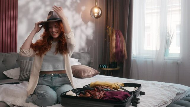 Happy Caucasian redhead ginger female positive smiling woman girl lady at home bedroom on bed preparing trip vacation travel holiday preparation clothes in suitcase baggage luggage put on summer hat