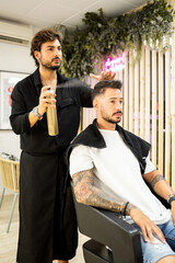 Vertical photo of an adult hairdresser with a beard and black clothes is using a can of spray...
