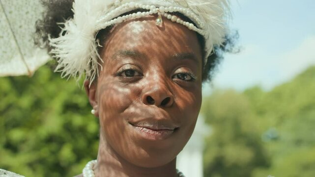 Portrait of young African American lady wearing feathered fascinator standing under lace parasol outdoors on sunny day, looking at camera and happily smiling