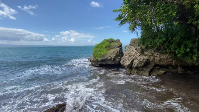 4k video footage of Hidden rocky beach with cliffs at Batukaras, Pangandaran, West Java, Indonesia during sunny day. Wavy beach with blue sky and clouds background.