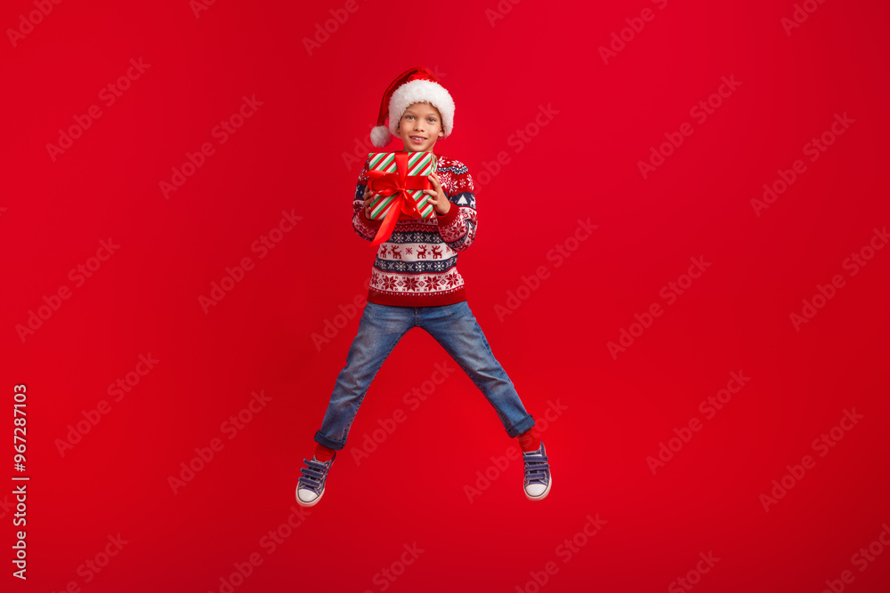 Wall mural Full length photo of charming small boy jumping hold gift celebrate christmas dressed knitted sweater isolated on red color background