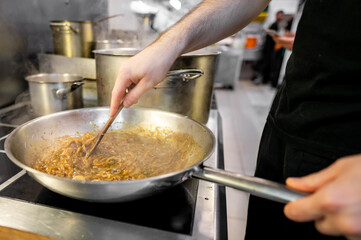 A close-up of a person stirring food in a pan on a stove, with pots in the background, capturing the action of cooking in a kitchen. Ideal for culinary, home cooking, and lifestyle themes.