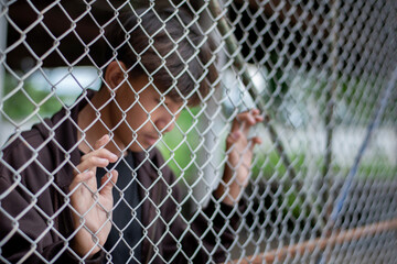 Asian schoolboy standing and holding a metal fence in local school detention center, looking down...