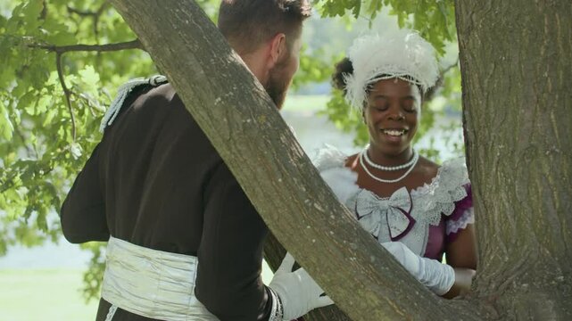 African American aristocratic lady hiding behind tree and then covering face with vintage hand fan while flirting with Caucasian officer on romantic date in park