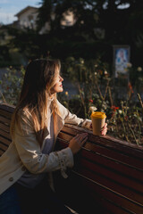 Woman drinks from cup on wooden bench. She is wearing a white shirt enjoying her beverage. The bench is located in a park setting, with trees in the background.