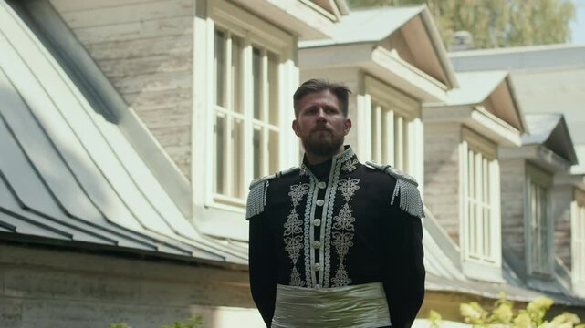 Caucasian man dressed in ornate military-style uniform of 19th century walking outdoors along house on summer day