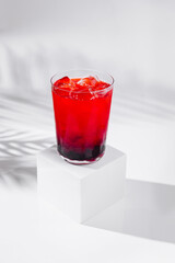 Red cocktail with ice and blackberries on white background with palm leaf shadow