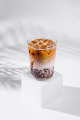 Iced coffee latte with chocolate syrup standing on white cube with tropical leaf shadow