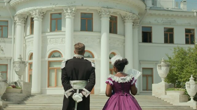 Tilt down shot of diverse aristocratic couple in bygone era costumes walking to royal palace and having conversation on summer day outdoors