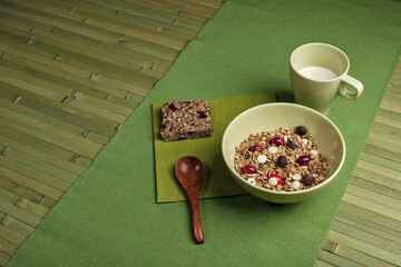 A healthy breakfast setup featuring a bowl of granola with dried fruits and white chocolate chips, a granola bar, and a cup of milk. The scene is arranged on a green placemat.