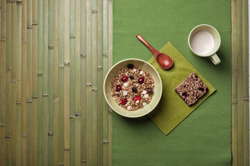 A simple and healthy breakfast arrangement featuring a bowl of granola with dried fruits and white chocolate chips, a granola bar, and a cup of milk on a green placemat with a wooden spoon.