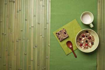 A minimalistic breakfast setting with a bowl of granola topped with dried fruits and white chocolate, a granola bar, and a cup of milk, placed on a green placemat with a wooden spoon.