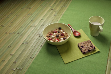 A cozy breakfast with a bowl of granola, dried fruits, white chocolate chips, a wooden spoon, granola bar, and cup of milk, all arranged on a green placemat and bamboo mat beneath.