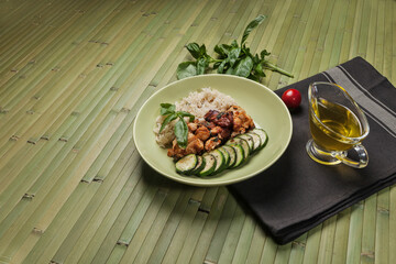 A healthy meal of grilled chicken, brown rice, and zucchini slices, garnished with basil. Served on a green plate with olive oil, tomato, and spinach leaves on a bamboo mat.