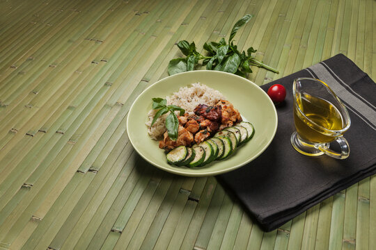 A healthy meal of grilled chicken, brown rice, and zucchini slices, garnished with basil. Served on a green plate with olive oil, tomato, and spinach leaves on a bamboo mat.