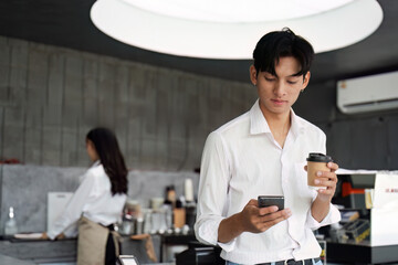 Young Man in Modern Cafe Using Smartphone and Holding Coffee Cup with Barista in Background