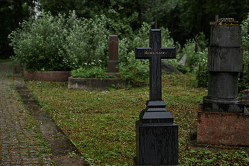 Background shot of rain wet memorial cross serving as lasting memorial to passed away standing in gloomy cemetery. Black tombstone with engraved epitaph reads Ruhe Shaft, copy space