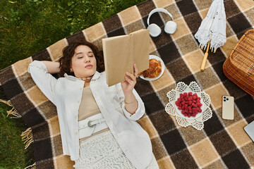 A pretty young woman relaxes on a blanket, savoring summer while reading a book during a picnic.
