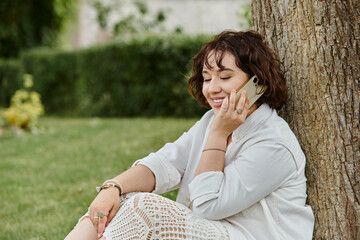 A young woman enjoys a sunny summer day, sitting against a tree and chatting on her phone in a lush green park.