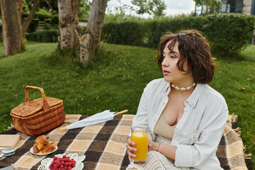 A young woman enjoys a peaceful moment, sipping juice while resting on a picnic blanket surrounded...