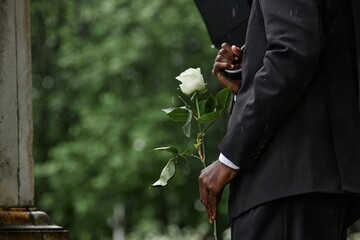 Cropped shot of Black man in funeral black suit holding white rose in memory of deceased loved one...