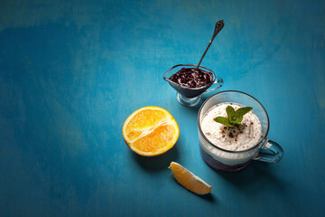 A delightful dessert arrangement with a glass of whipped cream layered with blueberry compote, garnished with mint, served alongside a halved orange and a small dish of blueberry jam on a blue table.