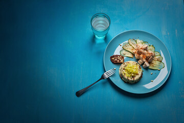 A vibrant meal on a blue plate featuring couscous, grilled chicken, cucumber slices, a stuffed date with pine nuts, and a glass of water. The presentation is set on a bright blue table.
