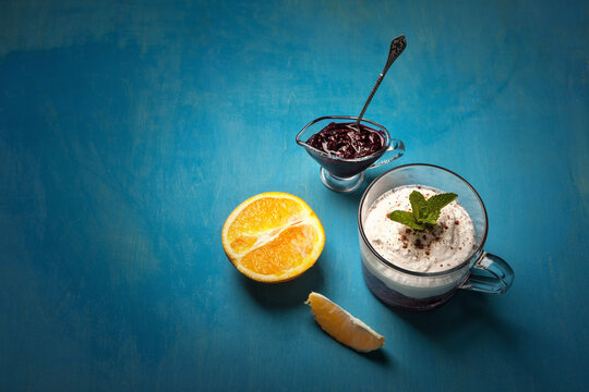 A delightful dessert arrangement with a glass of whipped cream layered with blueberry compote, garnished with mint, served alongside a halved orange and a small dish of blueberry jam on a blue table.