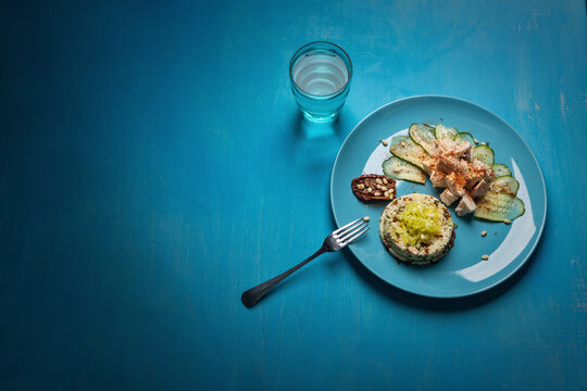 A vibrant meal on a blue plate featuring couscous, grilled chicken, cucumber slices, a stuffed date with pine nuts, and a glass of water. The presentation is set on a bright blue table.