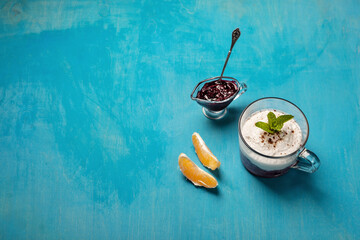 A light dessert featuring whipped cream topped with mint in a glass, blueberry jam in a small glass dish, and two orange slices on a bright blue background. The setup is minimal and refreshing.