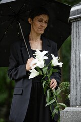 Vertical shot of senior woman in funeral suit holding white lilies visiting grave of departed loved...