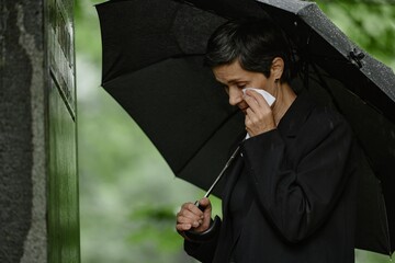 Side view of senior woman grieving next to memorial monument at grave wiping tears with handkerchief while hiding from rain under black umbrella at cemetery, copy space