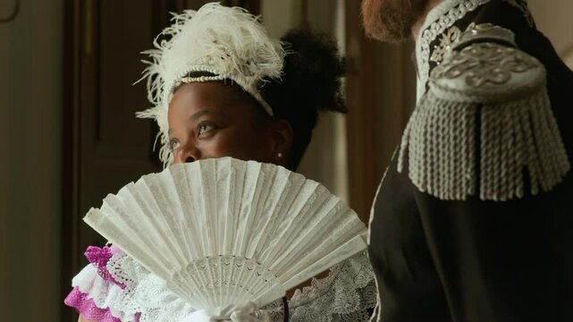 Young flirtatious Black woman in feathered fascinator covering face with vintage hand fan and talking with Caucasian officer standing beside in room