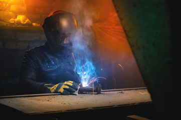 A welder in a leather jacket and welding mask welds in a dark room of an old barn. Home welding