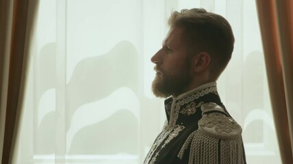 Tilt up side view shot of bearded officer in ornate period uniform with silver embroidery standing against window in living room