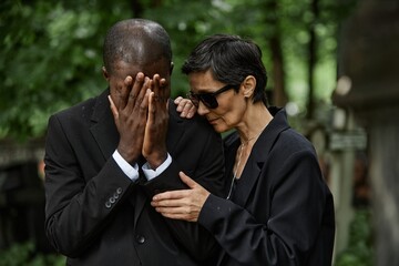 Medium shot of senior woman in sunglasses consoling crying husband at memorial ceremony....